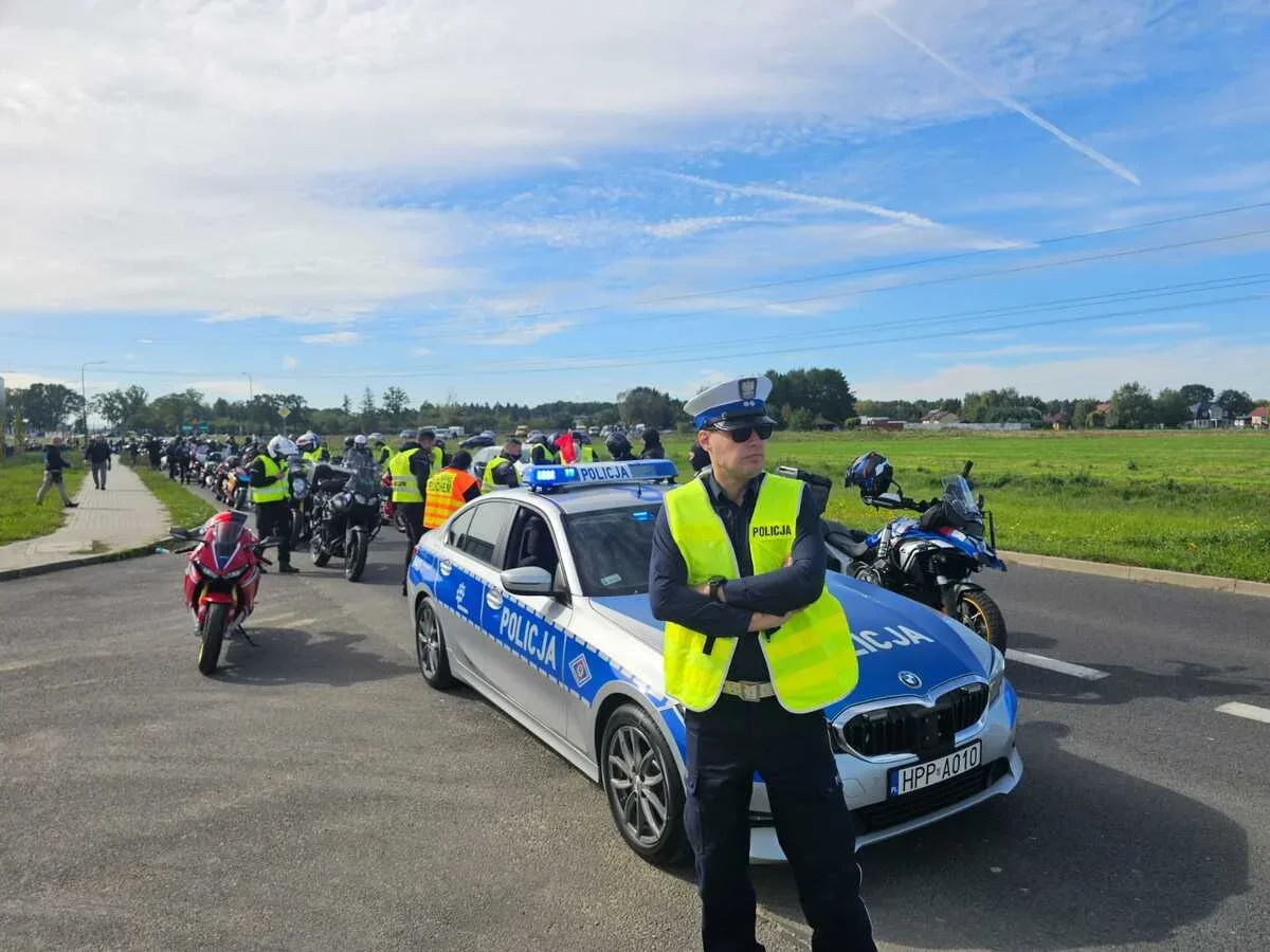 Policjant w kamizelce odblaskowej stoi przed radiowozem marki BMW oznaczonym „POLICJA” podczas zorganizowanej akcji z udziałem motocyklistów na wiejskiej drodze