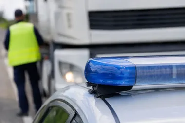 Policyjny radiowóz z niebieskim światłem na drodze, w tle funkcjonariusz w kamizelce odblaskowej przy ciężarówce.